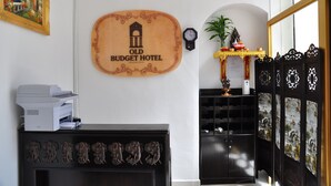 A reception area with a wooden sign reading "Old Budget Hotel," a clock, a printer, a decorative cabinet, and a potted plant. - Old Budget Hotel (George Town)