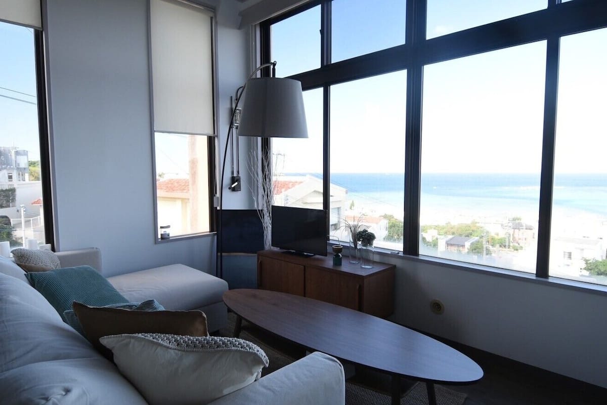 A modern living room with a sofa, a small table, a TV, and large windows offering a view of the sea.
