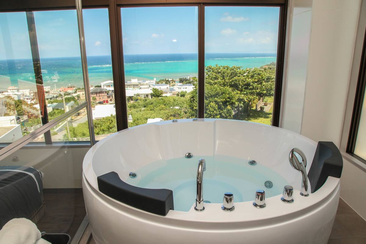 A large, round, white bathtub with multiple faucets, positioned in a room with large windows overlooking a coastal cityscape.