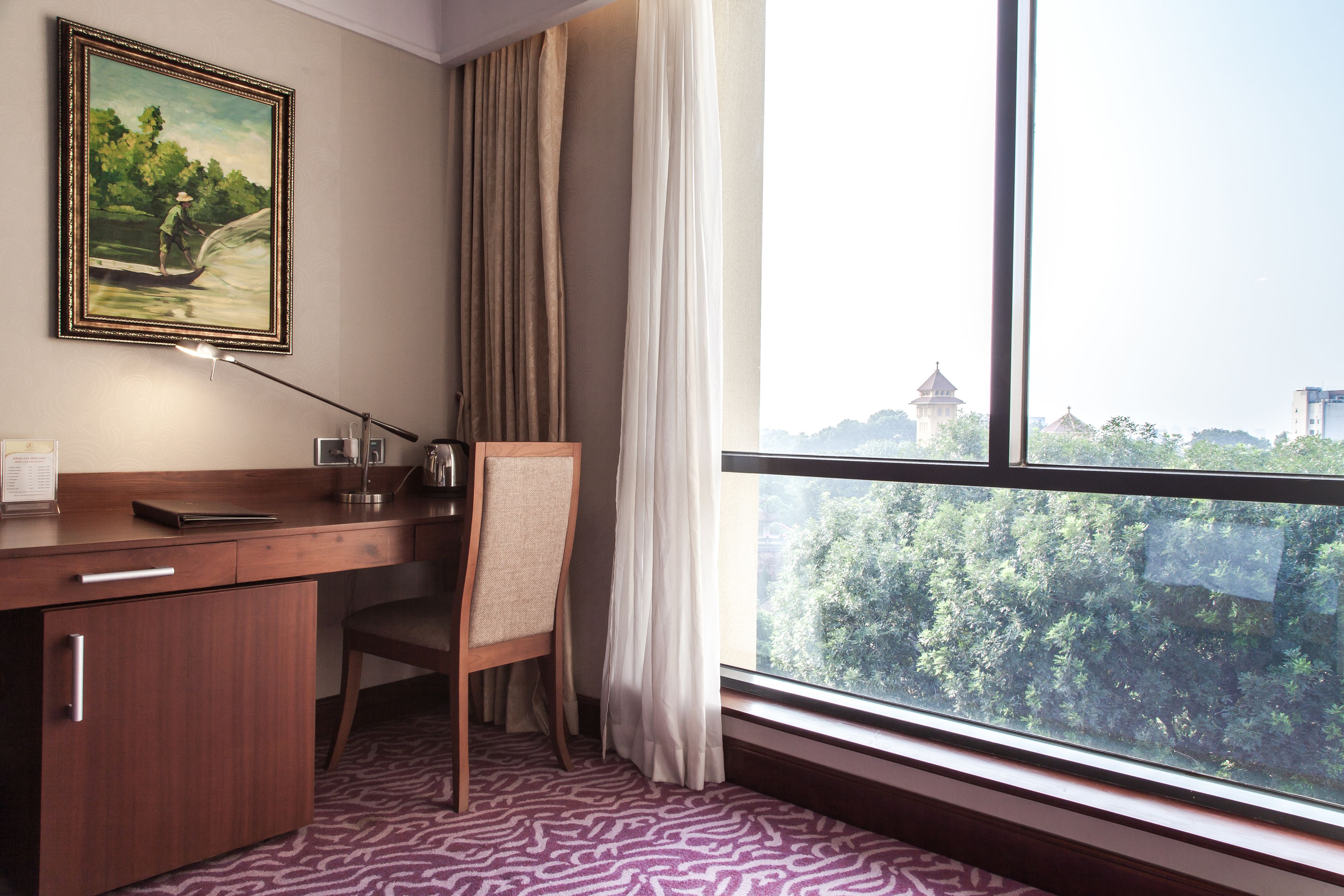 Habitación de hotel con escritorio, silla y un amplio ventanal que ofrece vistas a un paisaje urbano y áreas verdes.