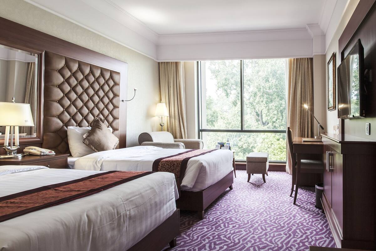 A hotel room with a large tufted headboard, two beds with white linens, a small desk, a chair, a window with a view of trees, and a television.