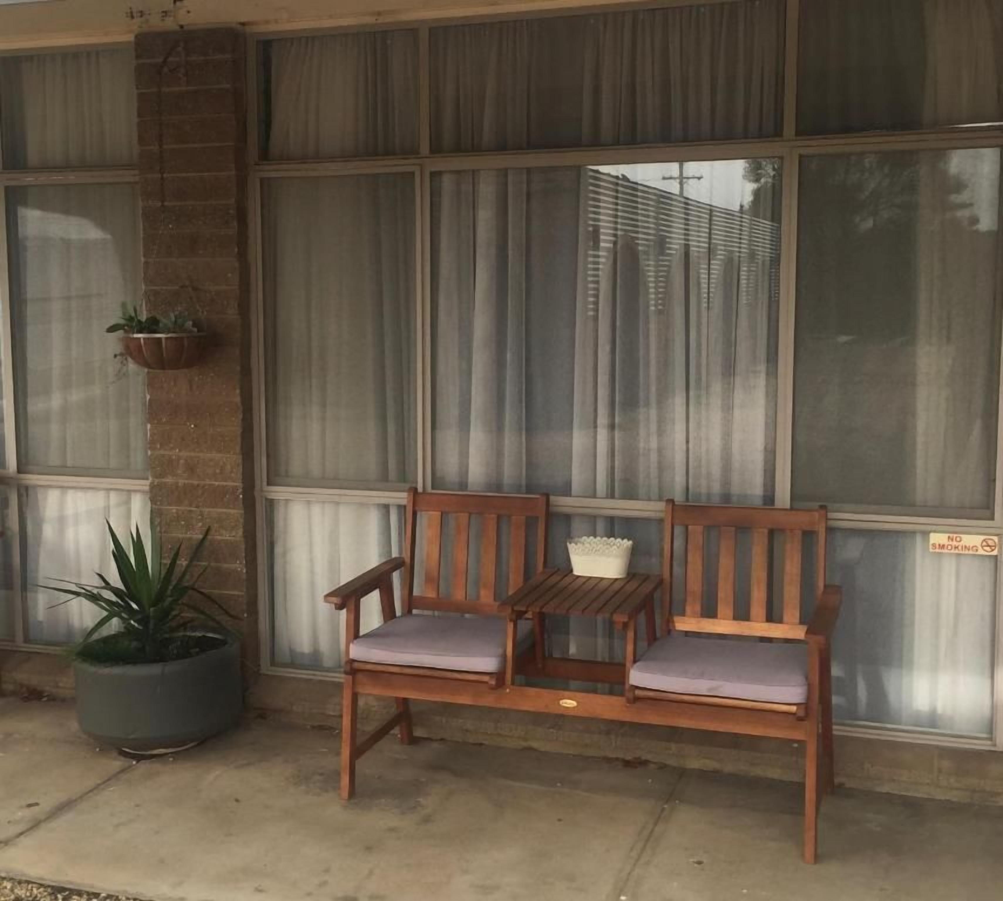 Un banco de madera con cojines frente a un edificio, una planta en maceta y una ventana con cortinas.