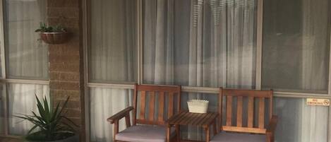 Un banco de madera con cojines frente a un edificio, una planta en maceta y una ventana con cortinas.