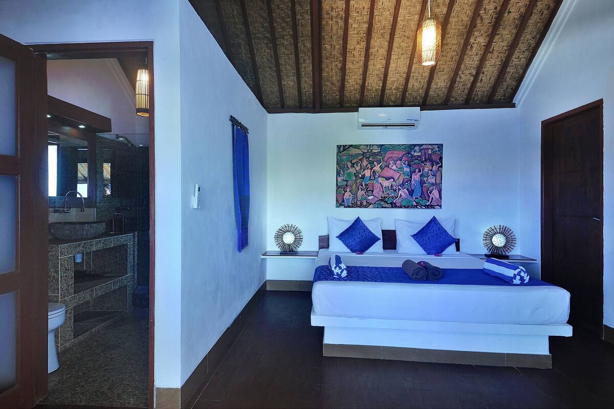 A bedroom with a bed, a nightstand, a wall-mounted air conditioner, a ceiling fan, and a large painting.
