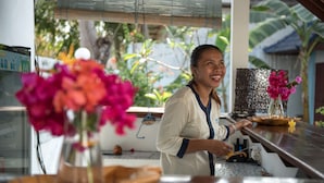 A person standing at a counter with a laptop, a vase with flowers, and a window with blinds in the background. - Marygio Resort (Gili Trawangan)