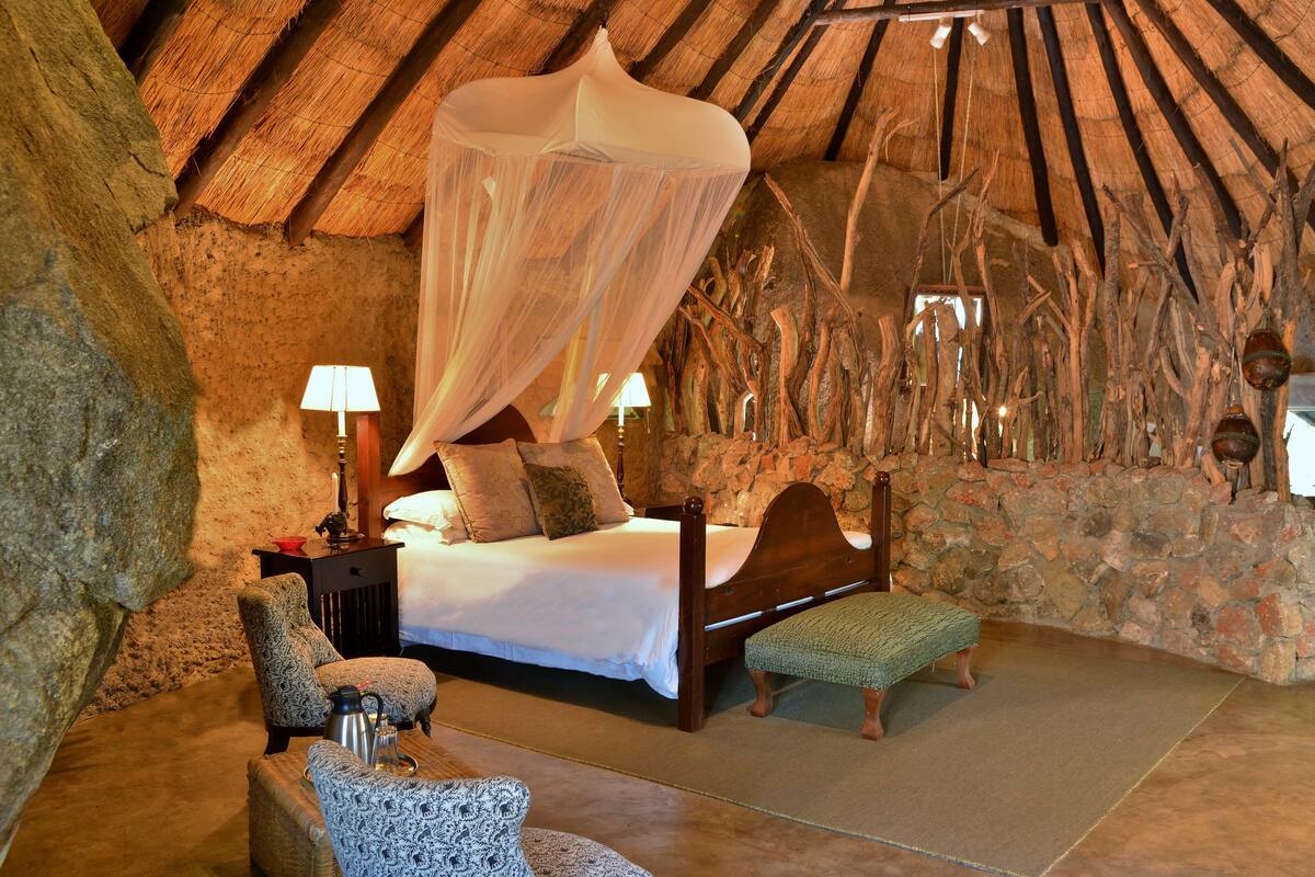 A rustic bedroom with a stone wall, a bed with a canopy, a nightstand with a lamp, and a chair.