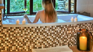 Une femme se détend dans un bain, entourée de bougies, avec un verre de vin à la main, et elle peut admirer la verdure à travers la fenêtre.