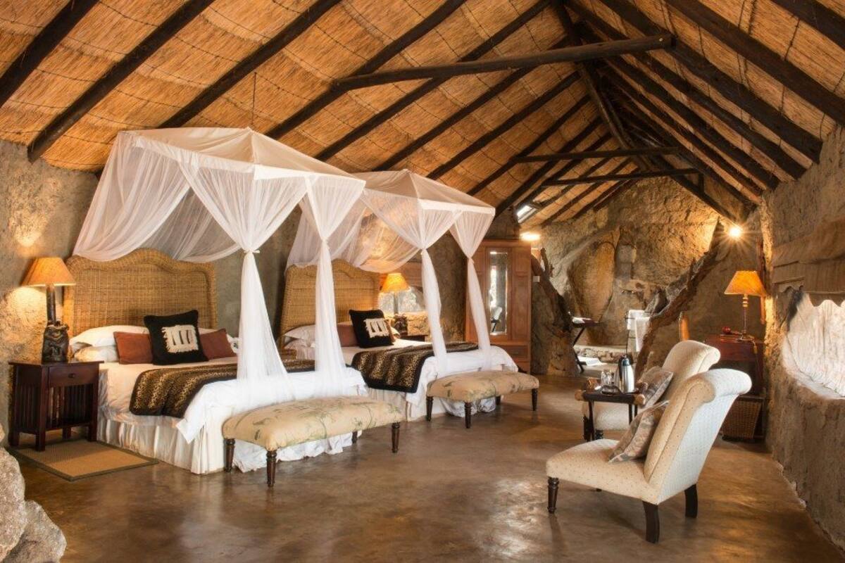 A rustic bedroom with four beds, each with a canopy, in a stone-walled room with a thatched roof.