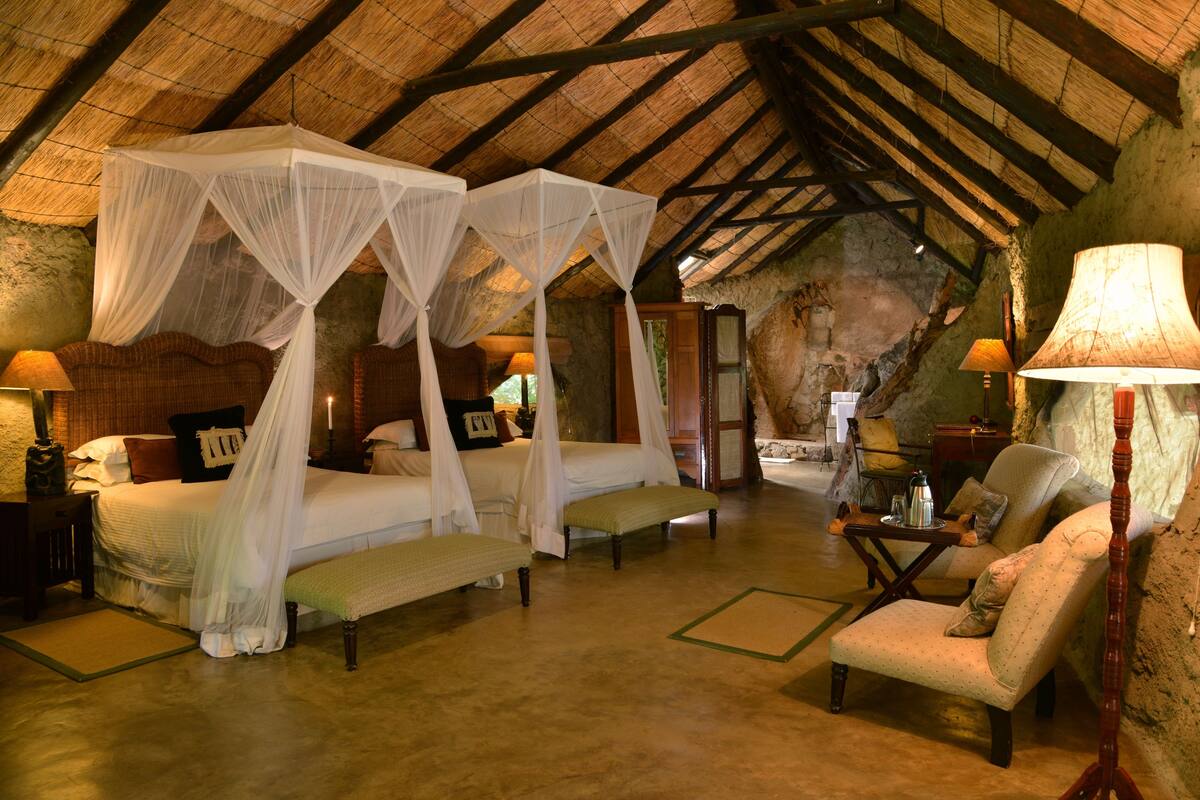 A room with a four-poster bed, a bench, a chair, and a table with a lamp.