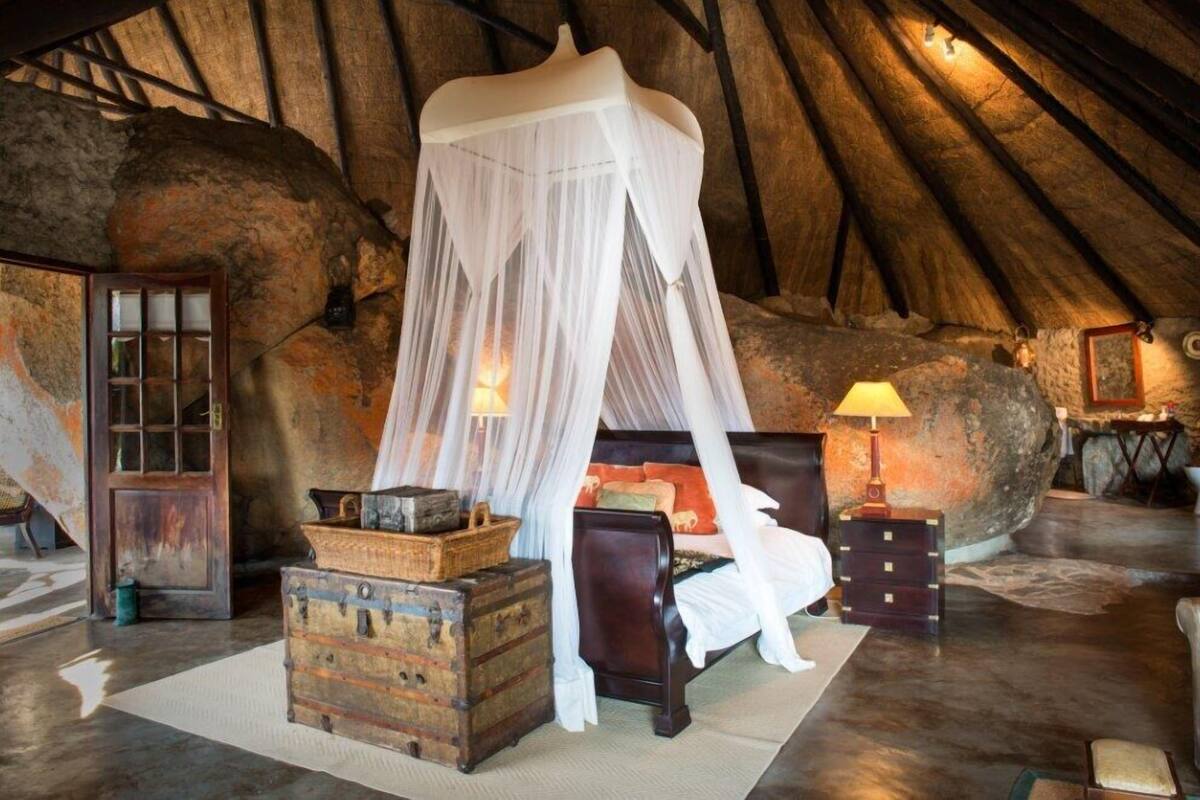A rustic bedroom with a four-poster bed, wooden trunks, and a stone wall backdrop.