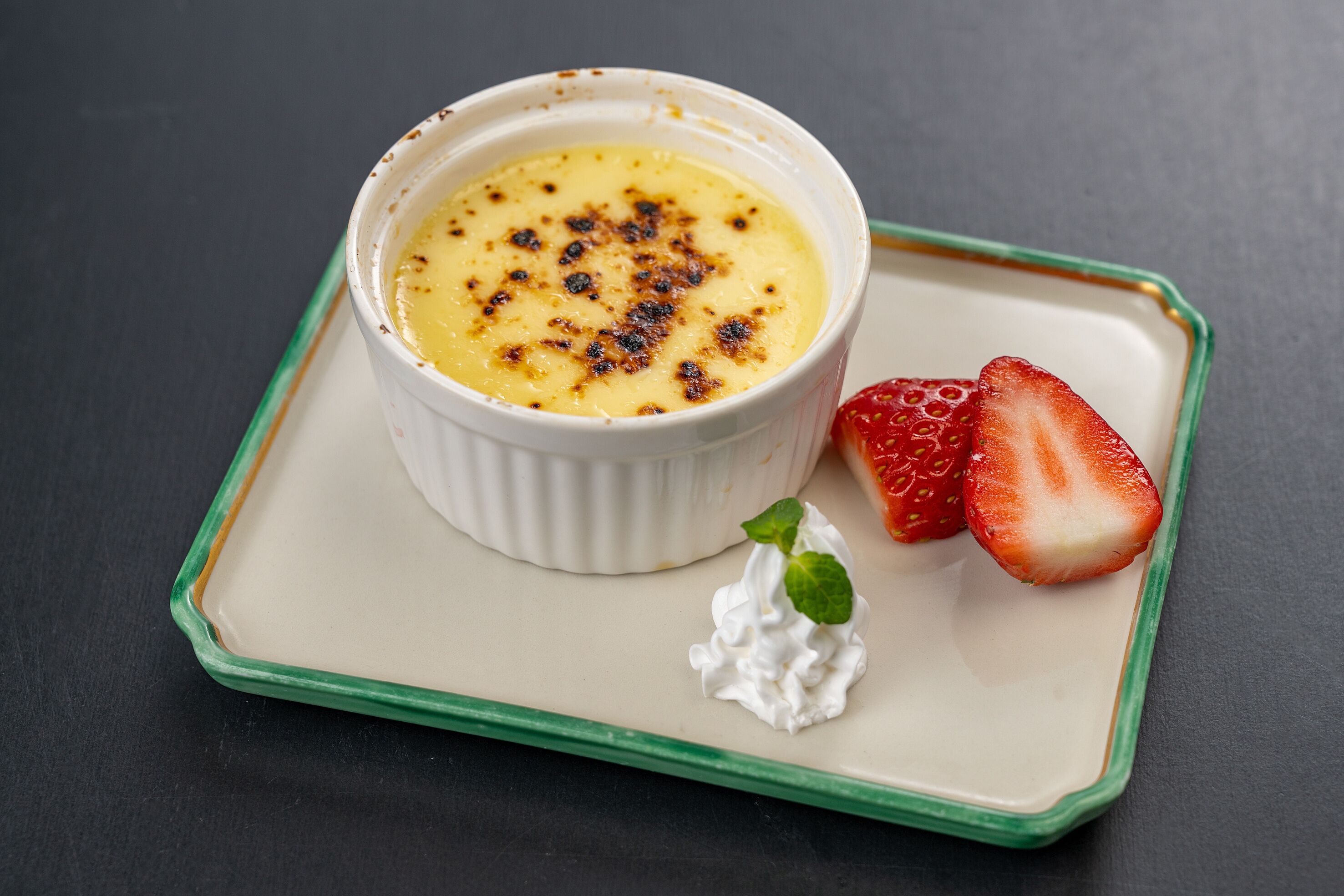 Un postre en un recipiente blanco sobre una bandeja verde, con dos fresas y una cucharada de crema batida.