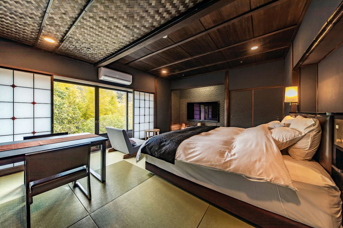A modern bedroom with a large bed, a desk, and a view of trees through large windows.