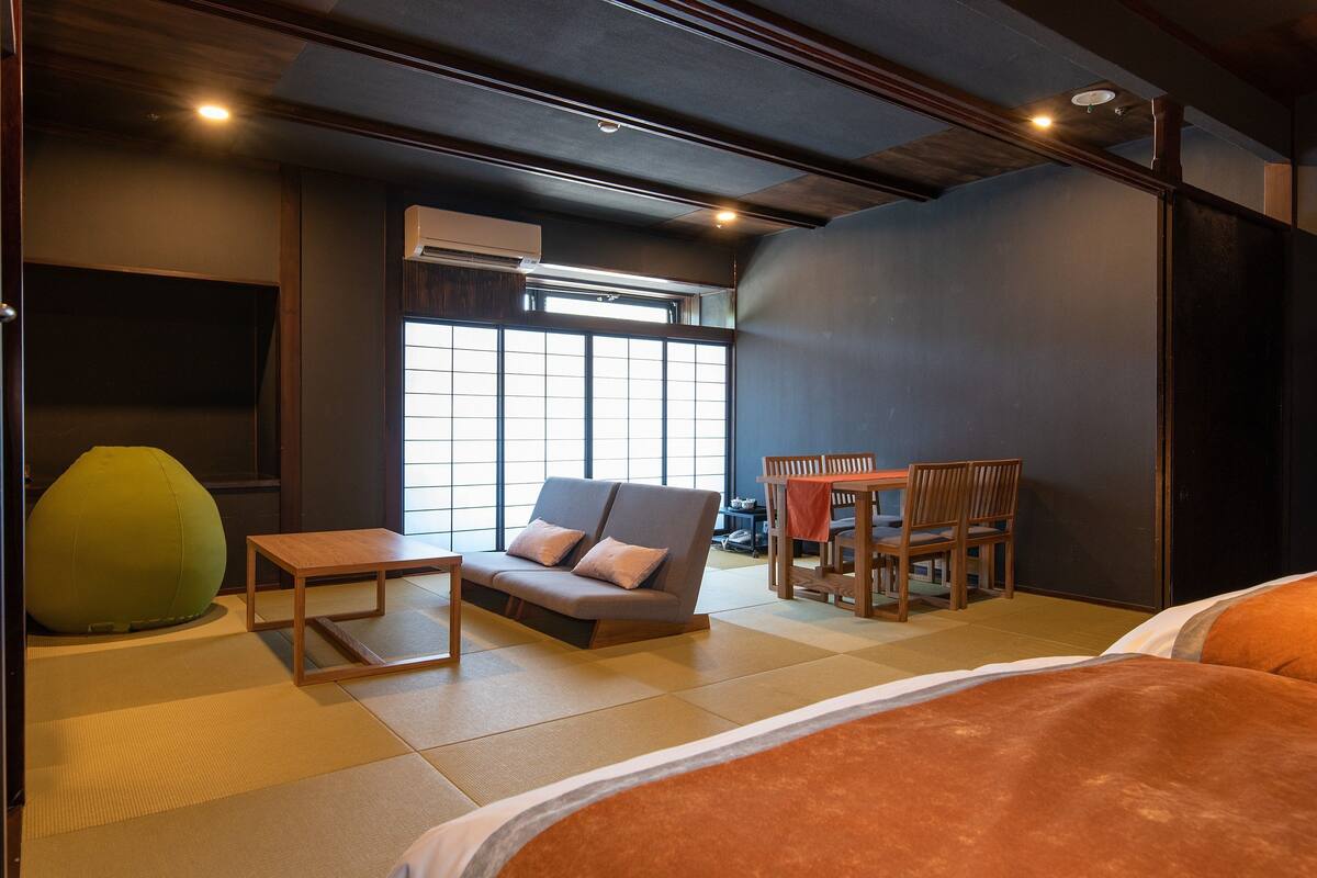 A modern hotel room with a sofa, a small table, a green bean bag, a dining area with chairs, and a large window providing natural light.