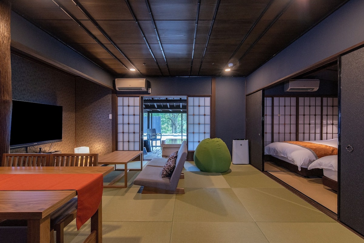 A traditional Japanese-style room with tatami flooring, a tatami mat table, a low wooden bench, a green bean bag, a bed with white bedding, and a television on the left wall.