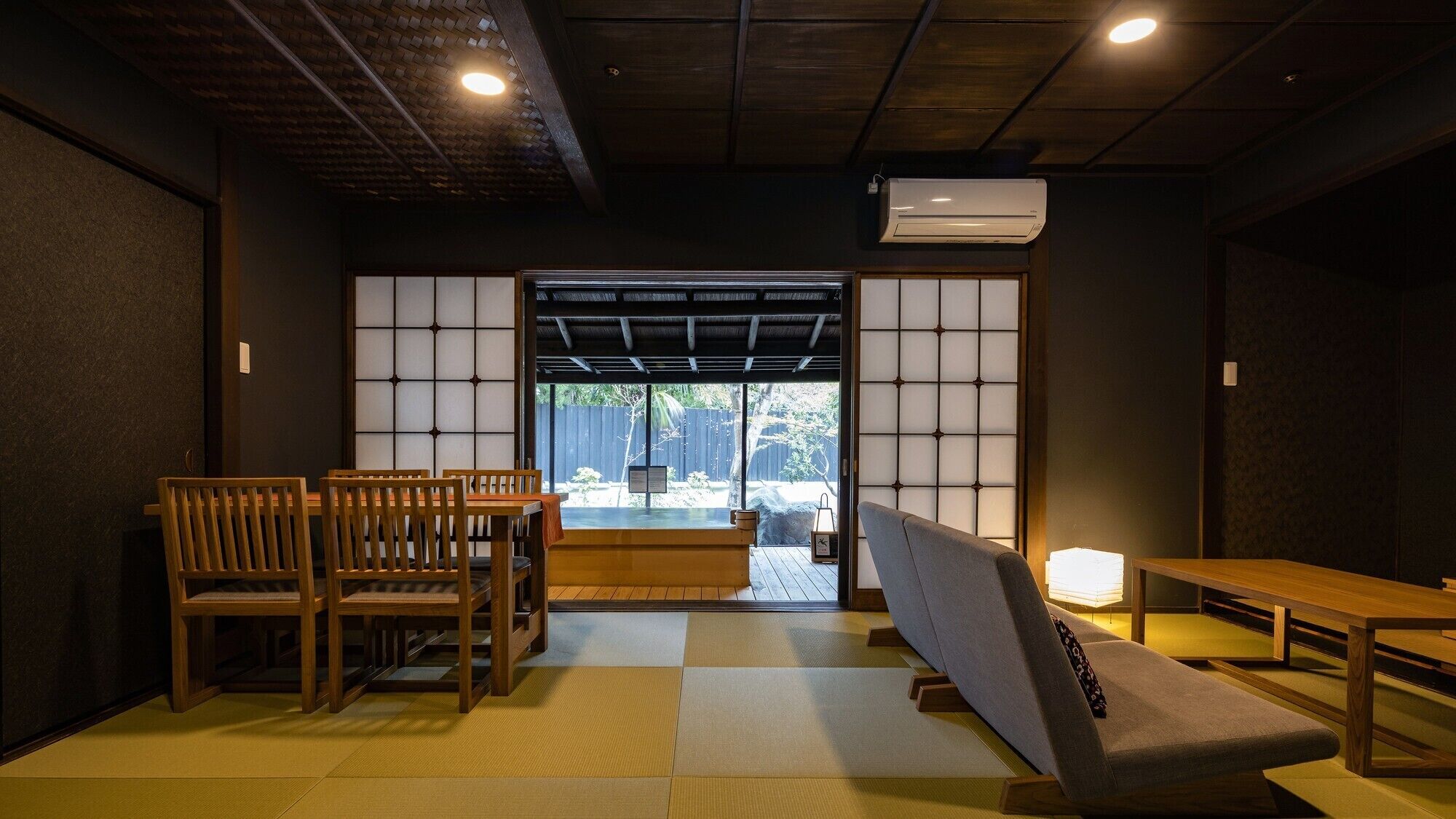 A traditional Japanese room with tatami flooring, a low table, wooden chairs, a sofa, and a sliding door leading to an outdoor area.