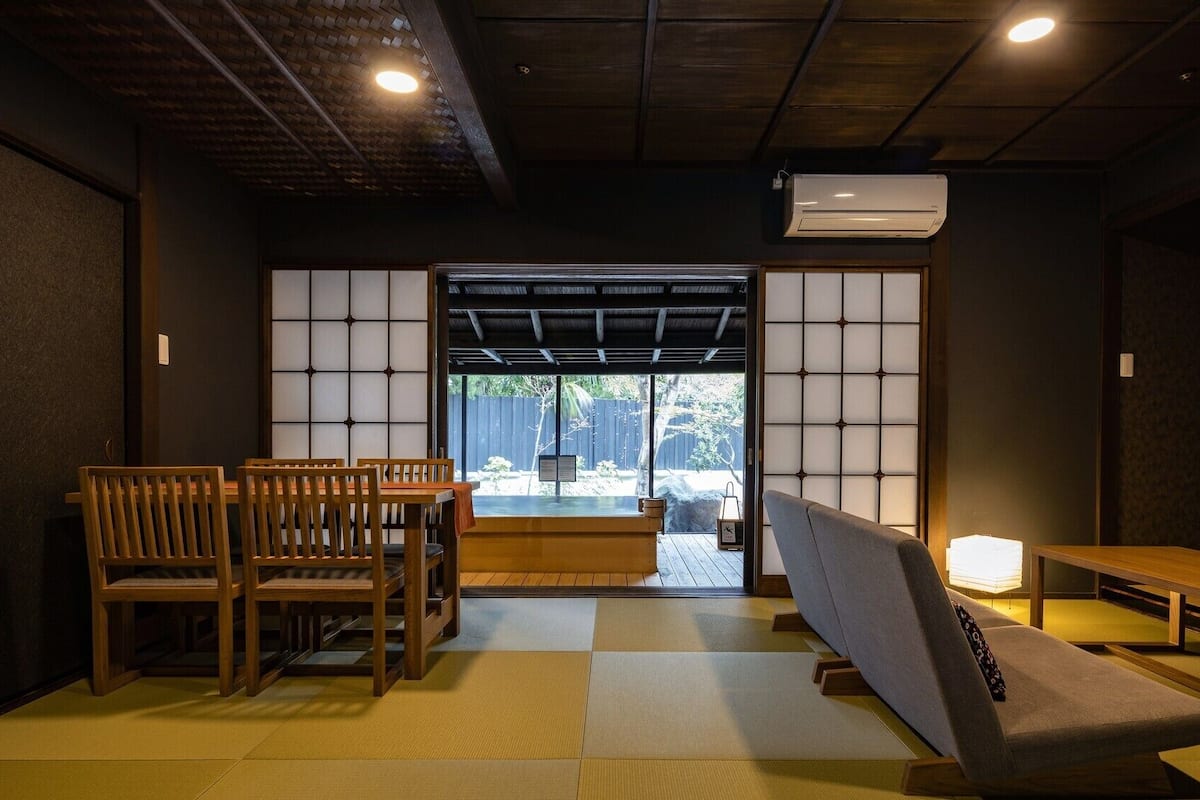 A traditional Japanese room with tatami flooring, a low table, wooden chairs, a sofa, and a sliding door leading to an outdoor area.
