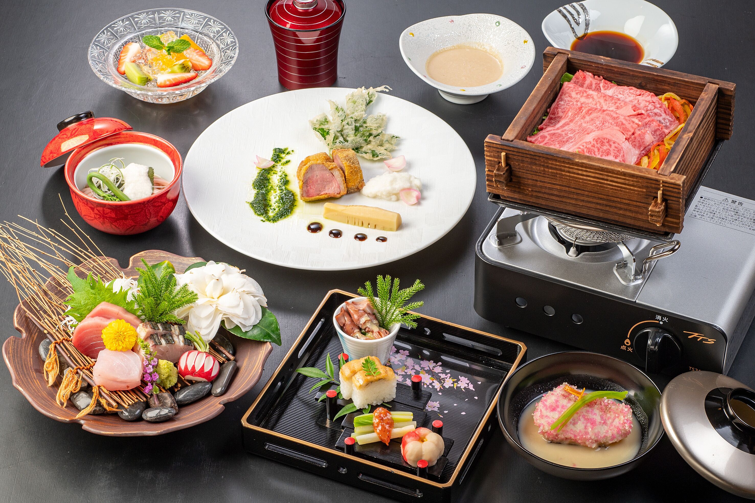 Variedad de platos japoneses, como sashimi, sushi y tempura, dispuestos sobre una mesa con una estufa a gas y un calentador de agua.