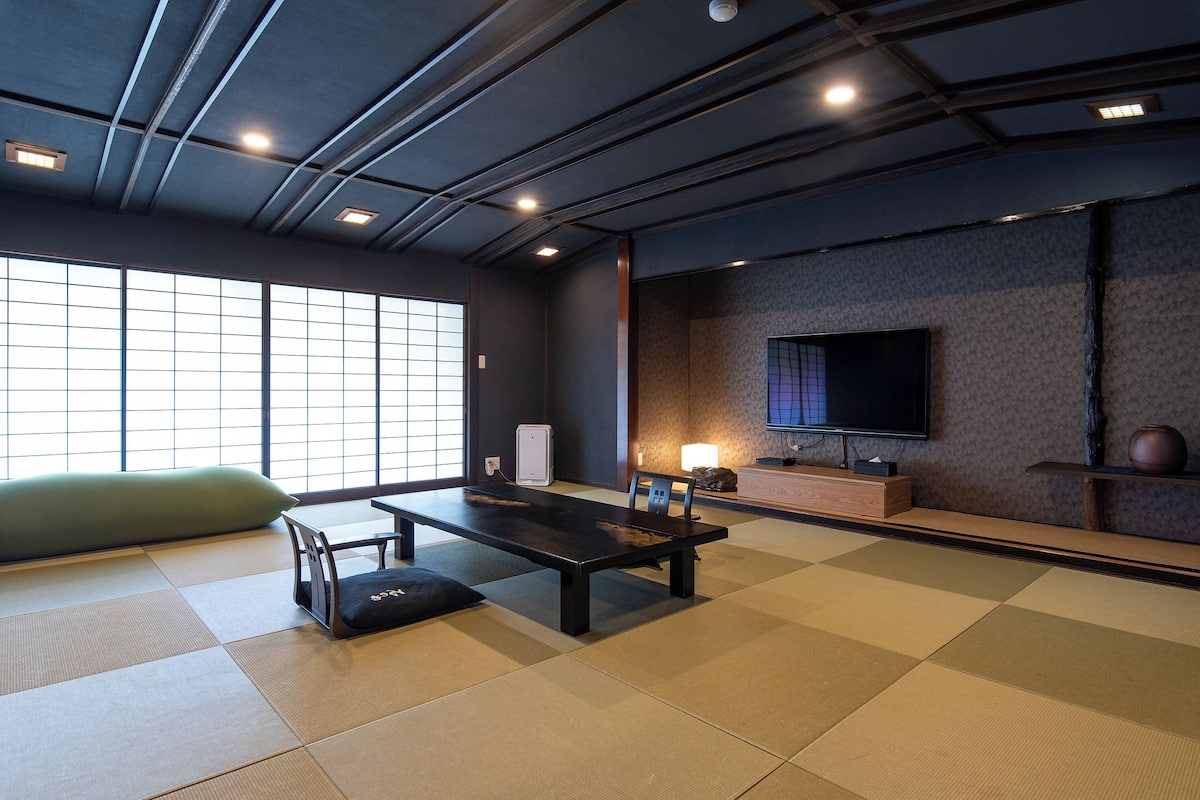 A tatami mat room with a low table, a green cushion, a TV, and a traditional Japanese cabinet.