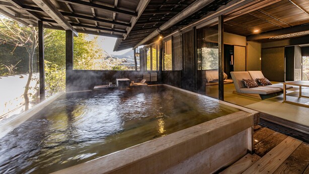 Zona de baño termal con vigas de madera, vista a árboles y un sofá con cojines.