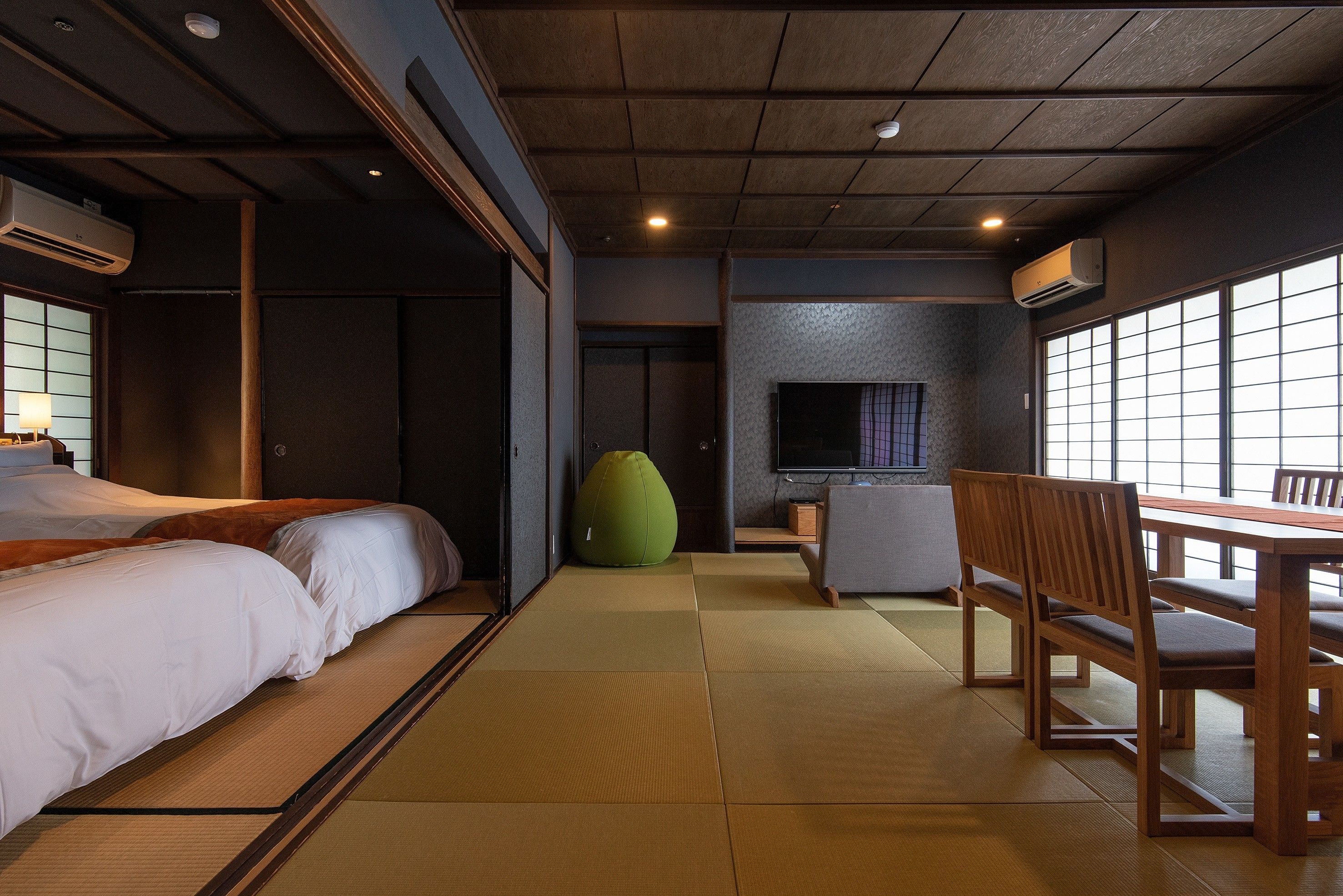 A room with tatami flooring, a bed, a green bean bag, a TV, and a dining table with chairs.