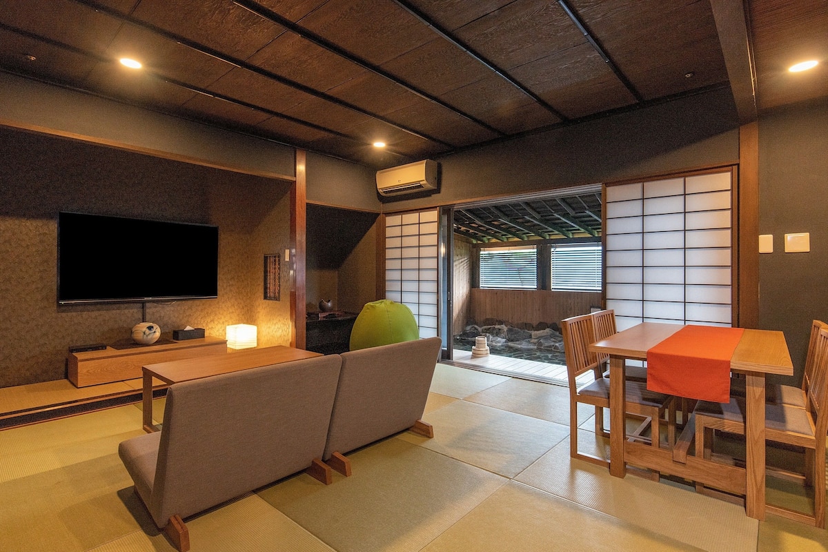 A traditional Japanese room with tatami flooring, a low wooden table with a red tablecloth, a sofa, a television, and a sliding shoji screen.