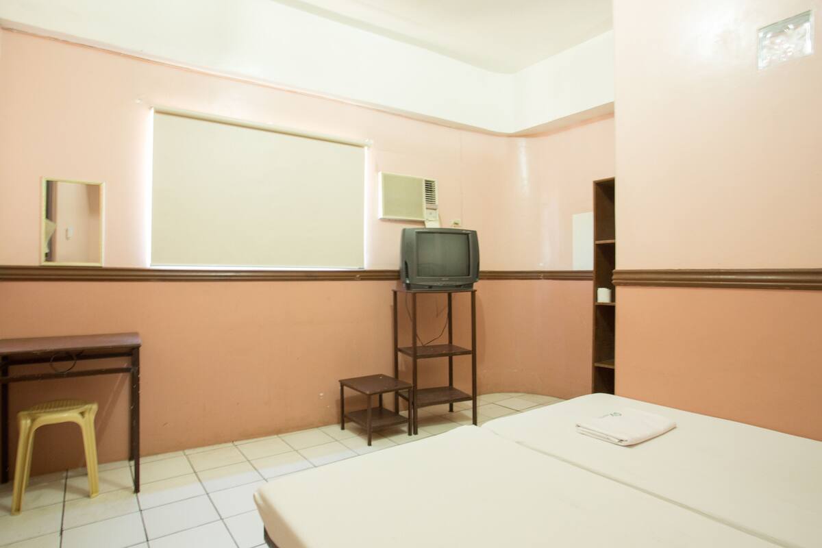 A small, simple room with a bed, a desk, a stool, a television on a stand, and a window.