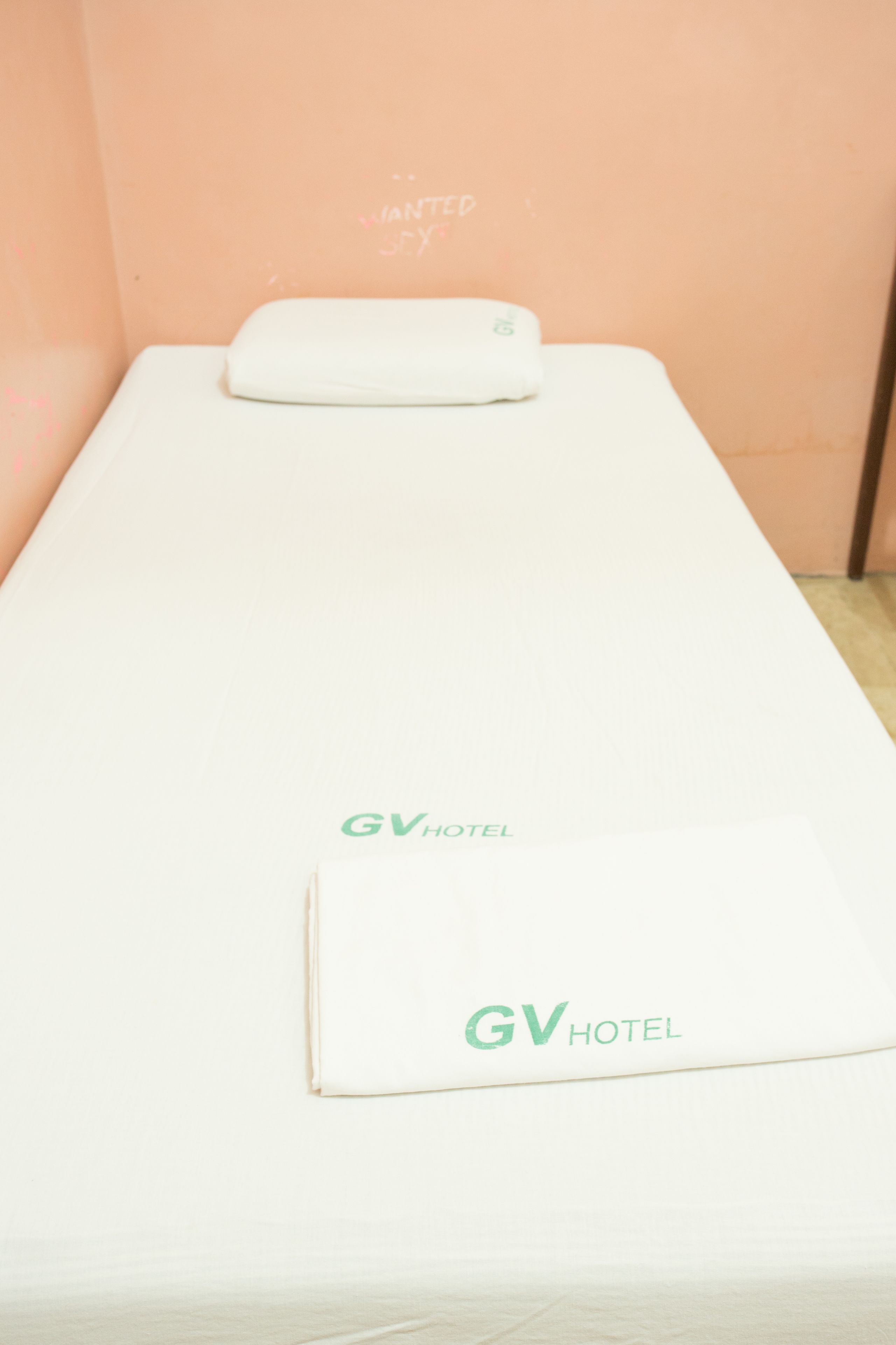 A single bed with a white mattress and a white pillow. The bed is placed against a peach-colored wall. The logo 'GV HOTEL' is visible on the mattress.
