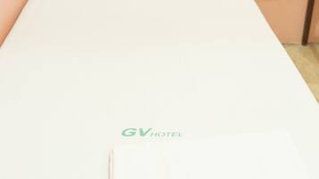 A single bed with a white mattress and a white pillow. The bed is placed against a peach-colored wall. The logo 'GV HOTEL' is visible on the mattress.