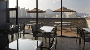 A rooftop dining area with tables and chairs, large windows, and a view of city buildings and mountains. - Hotel the May (Namyangju)