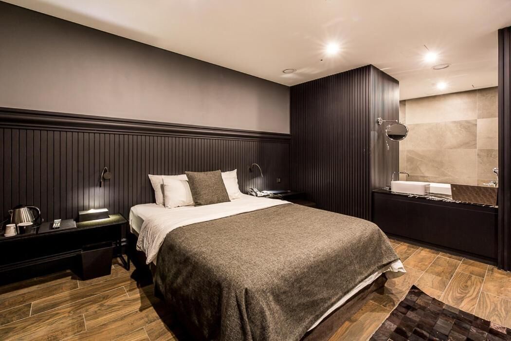Ein modernes Schlafzimmer mit einem großen Bett, dunklen Holzpaneelen, einem Nachttisch mit Lampe und einem Badezimmer mit Spiegel und Waschbecken.