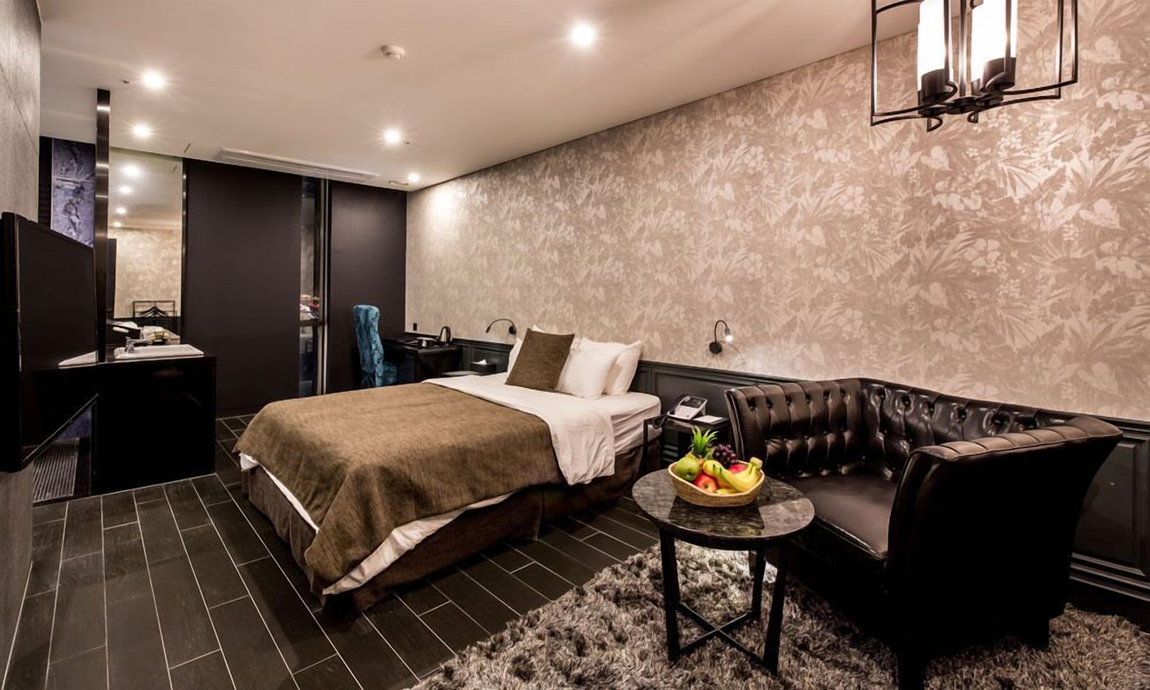 A modern hotel room with a large bed, a leather armchair, a small round table with fruit, and a decorative wall.