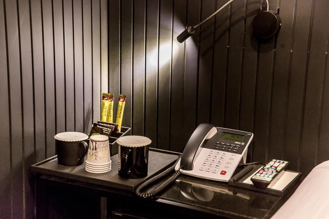 A hotel room with a bedside table featuring a telephone, a remote control, and a tray with coffee cups and a mug.