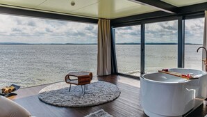 Una habitación moderna con un ventanal grande, un tapete redondo, una silla de madera, una bañera blanca y una mesa de madera.