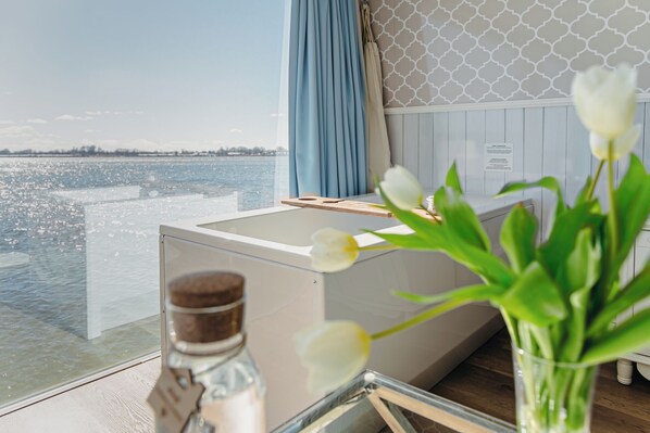 A room with a view of the water, a wooden table, a bottle, and a vase with flowers. - HT Houseboats (Mielno)