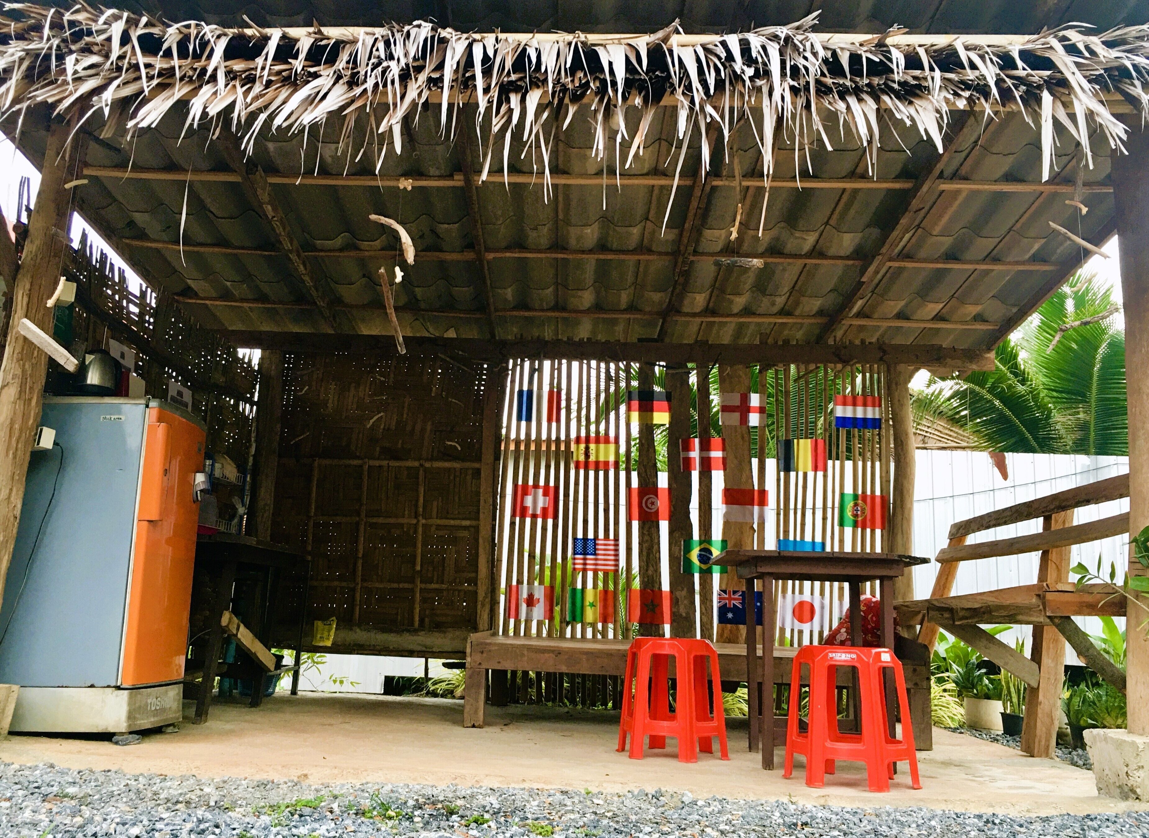 Un espacio al aire libre techado con paja, con un banco y taburetes de plástico rojo. Se exhiben banderas en la pared detrás del banco.
