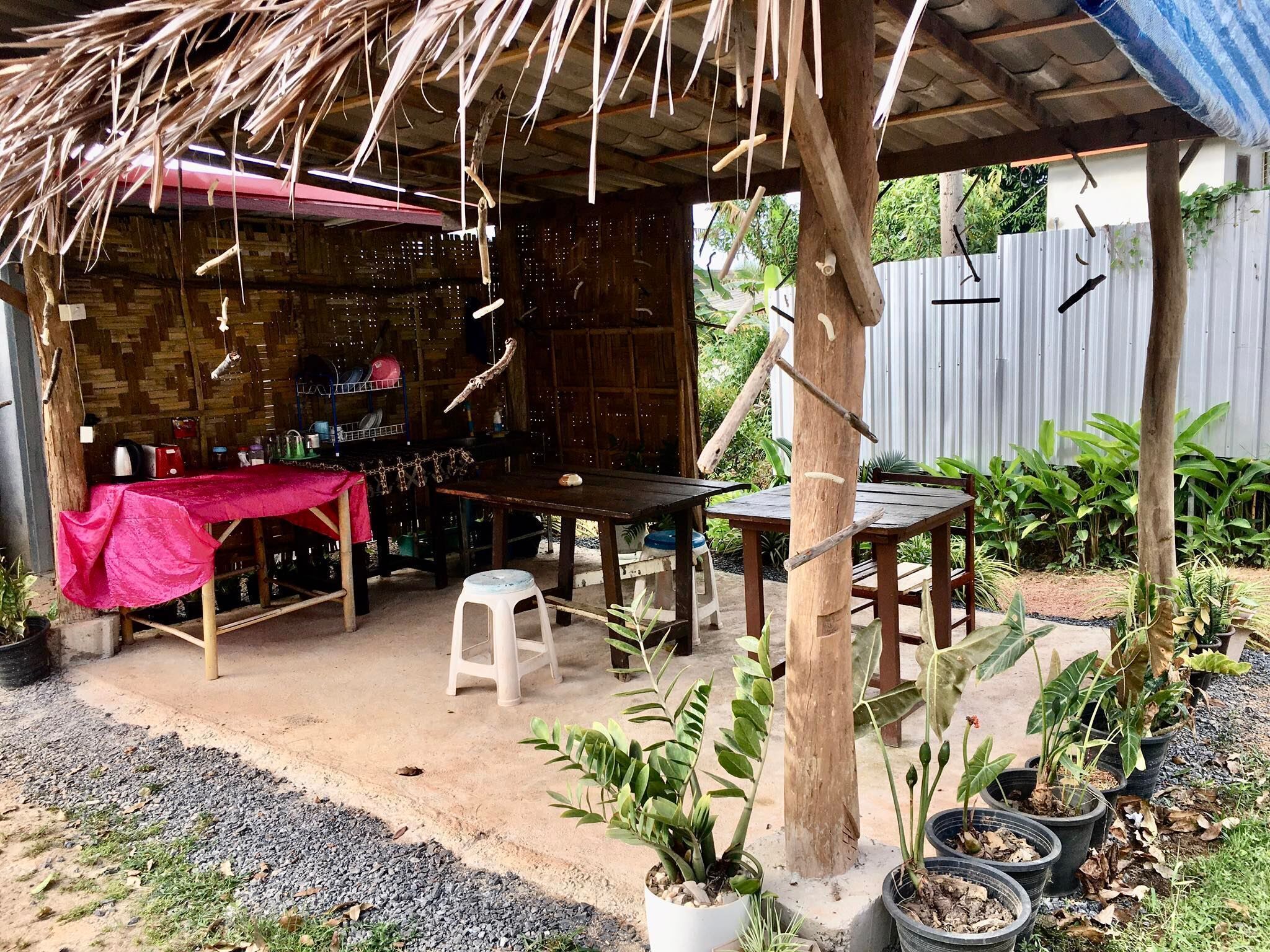 An outdoor patio area with a wooden table and chairs, a thatched roof, and potted plants.