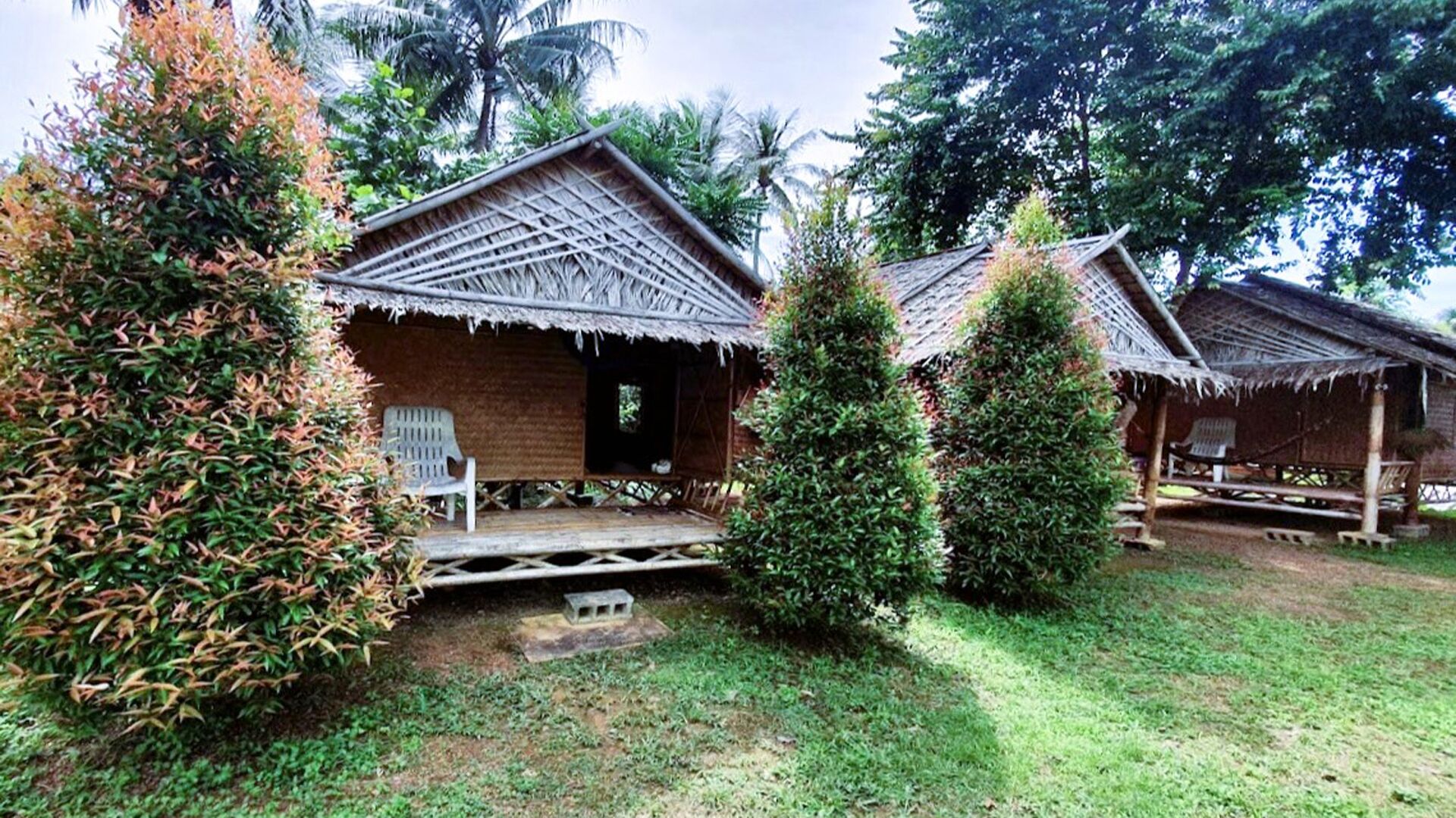 A rustic bungalow with a thatched roof, surrounded by lush greenery and palm trees.