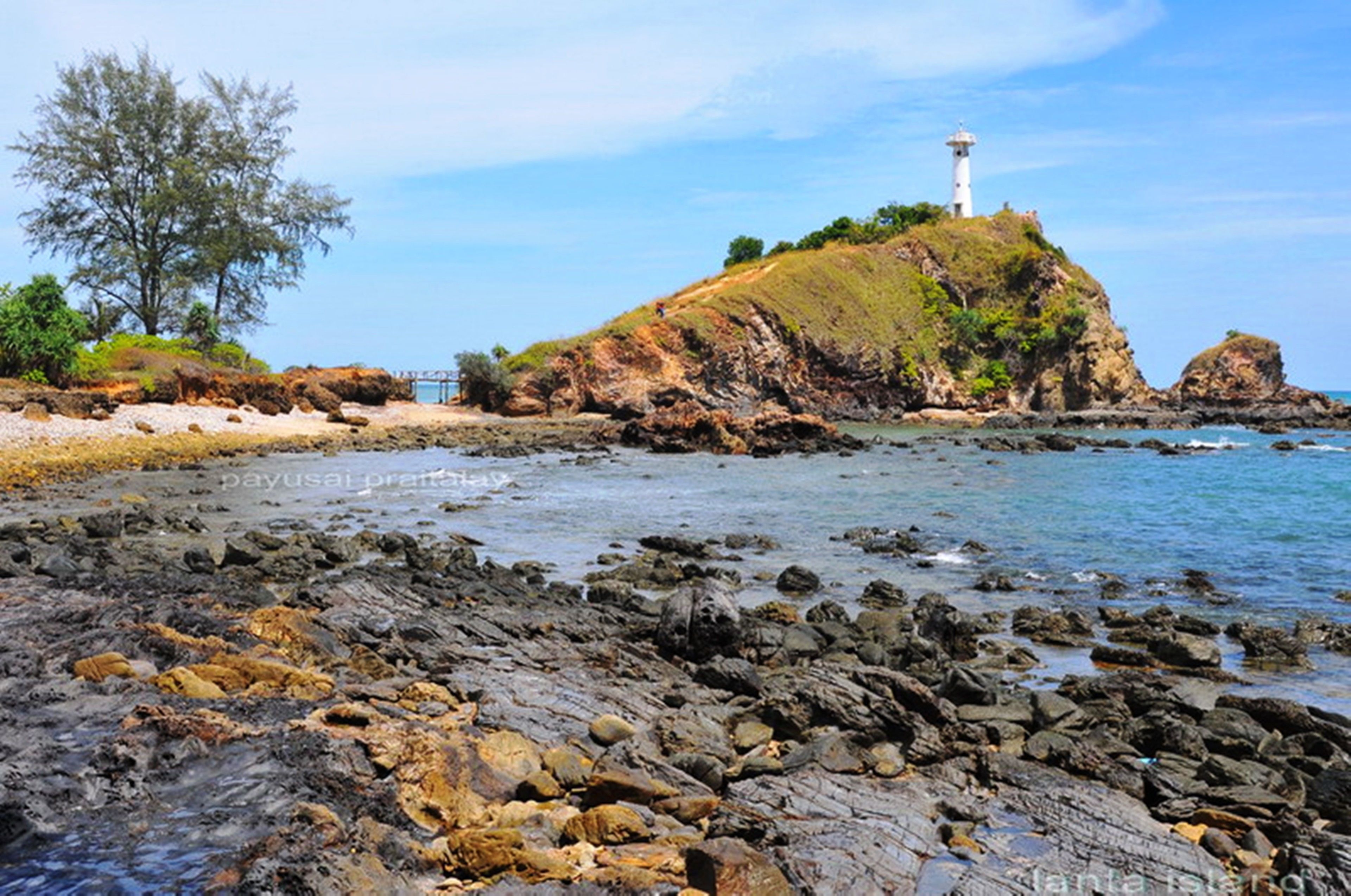 A rocky shoreline with a lighthouse on a hill, clear skies, and calm sea.