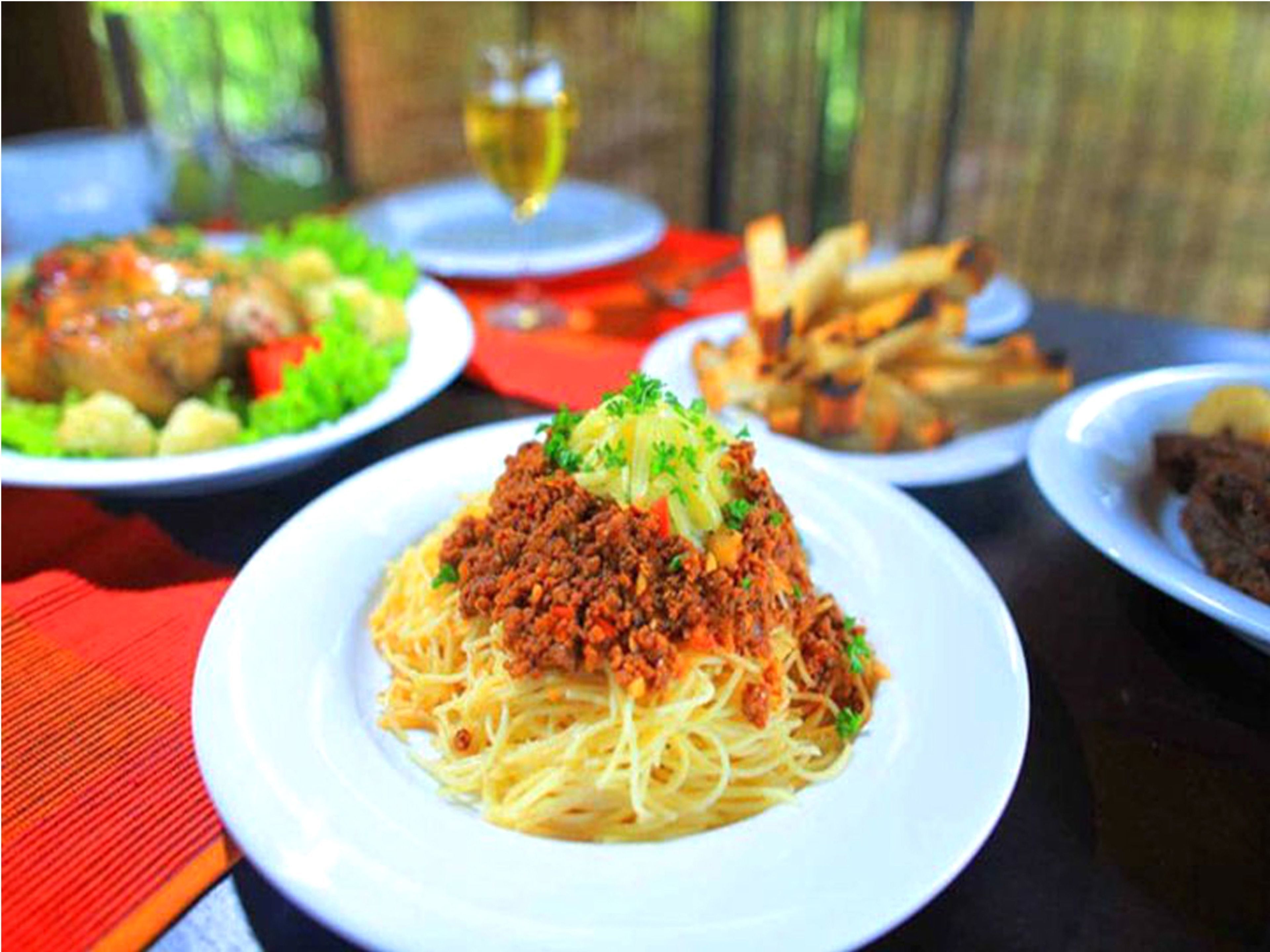A table with plates of spaghetti topped with meat sauce, a glass of white wine, and a plate of fries.