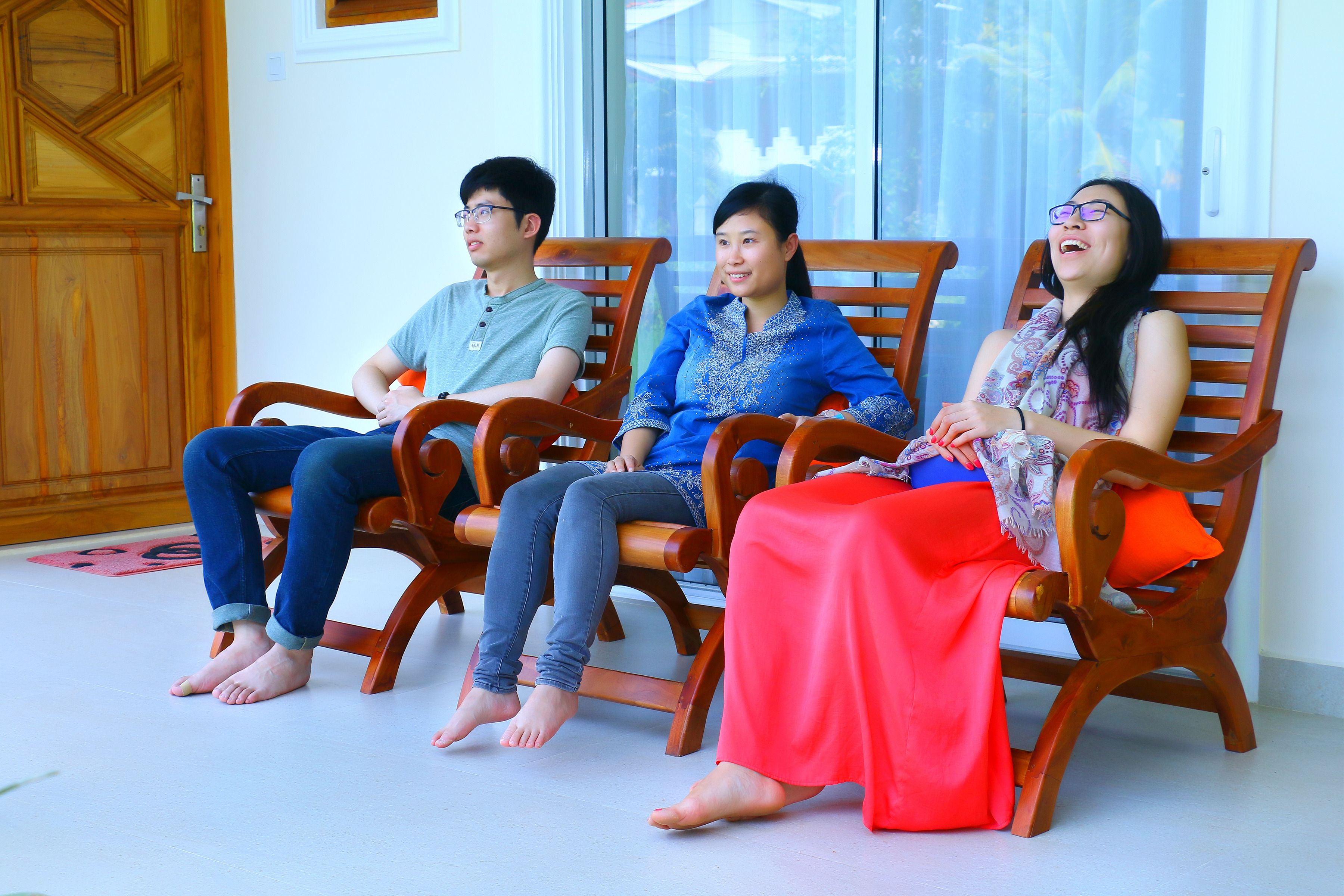 Three people sitting on wooden chairs in a room with a wooden door and a window with curtains.