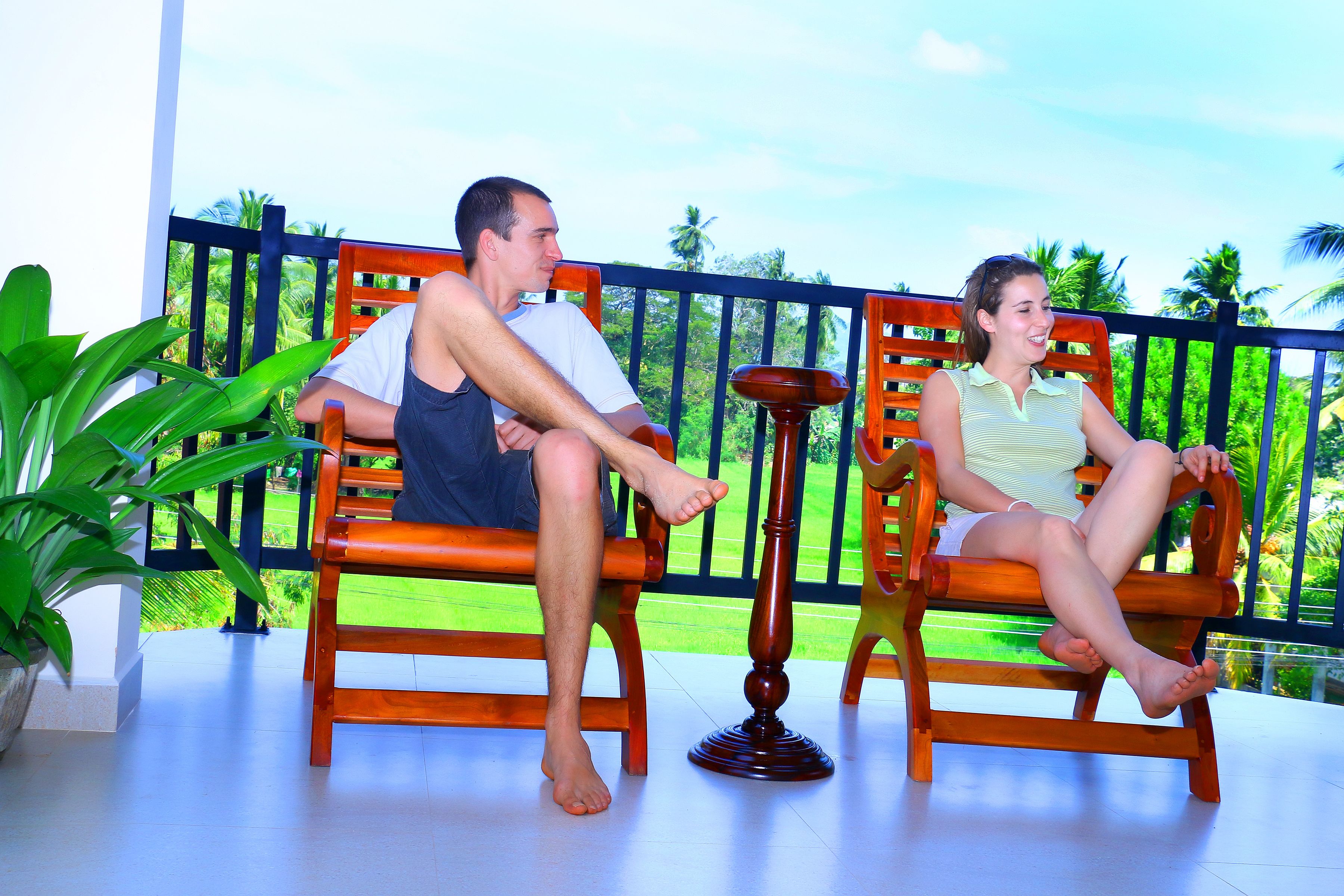 Two people sitting on wooden chairs on a balcony with a view of greenery and a fence.