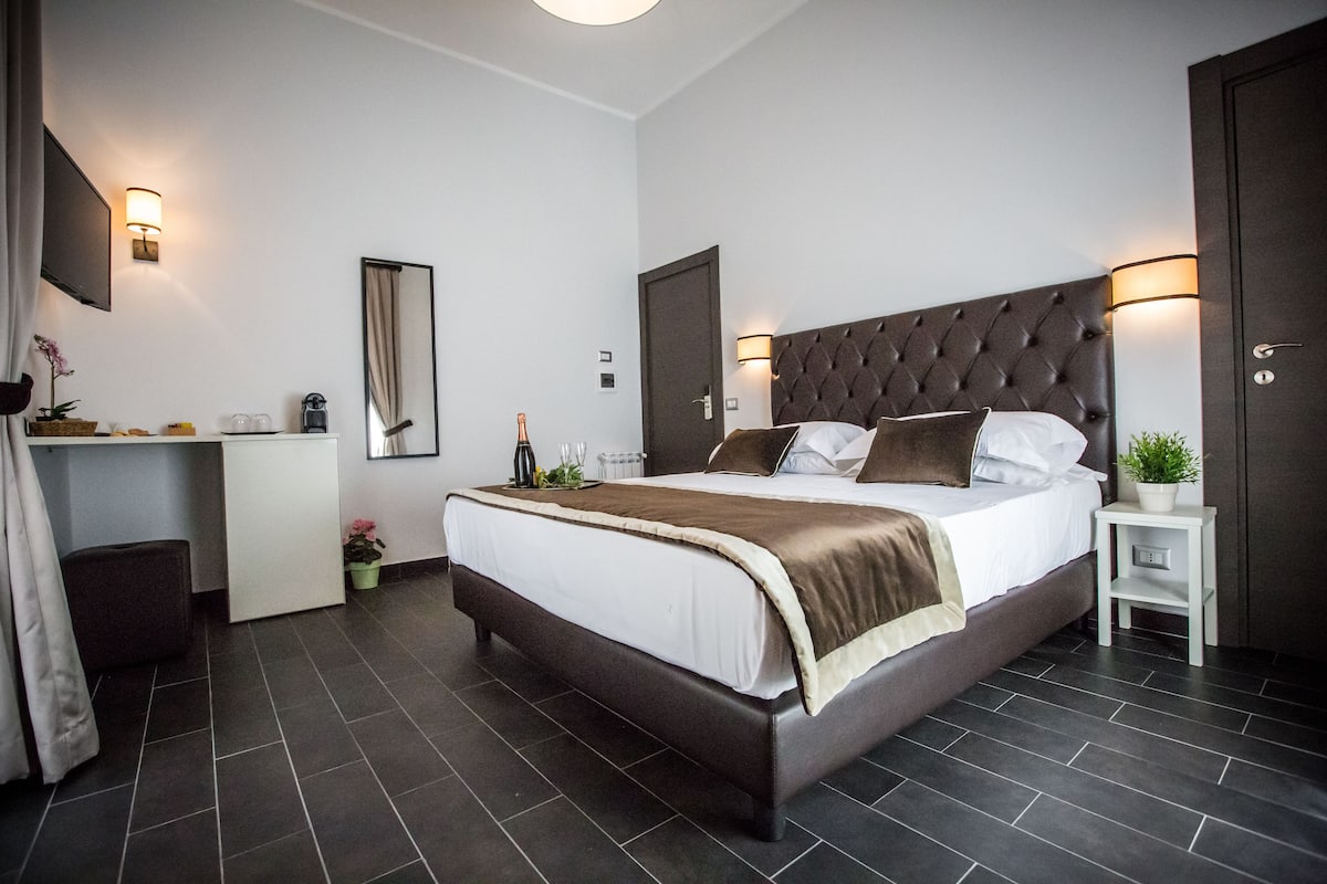 A modern hotel room with a large bed, a bedside table, a desk, and a mirror.