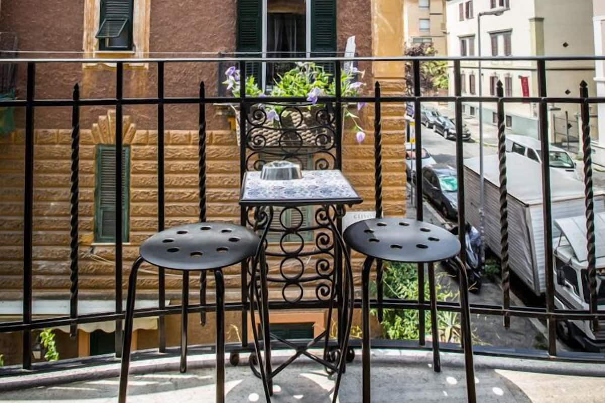 A balcony with two metal chairs and a small table, overlooking a street with parked cars and buildings.