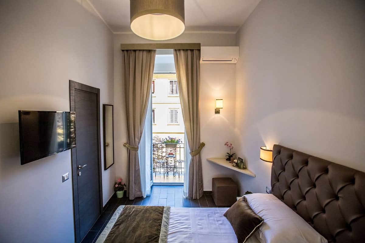 A hotel room with a bed, a TV mounted on the wall, a small table with a lamp, and a balcony with a view of buildings.