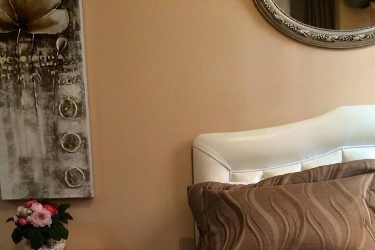 A neatly made bed with a striped bedspread and a rolled white towel. A white bedside table with a vase of flowers is visible.