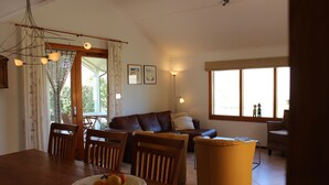 Ein Essbereich mit Holztisch, Stühlen, Obstschale, einer Couch und einem großen Fenster mit Vorhängen.
