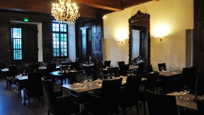 Ein schwach beleuchtetes Restaurant mit Holzbalken, Kronleuchtern und großen Fenstern. Die Tische sind mit Weingläsern und Tellern gedeckt.
