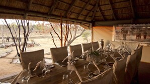 An outdoor dining area with a thatched roof, surrounded by trees and a view of wildlife.