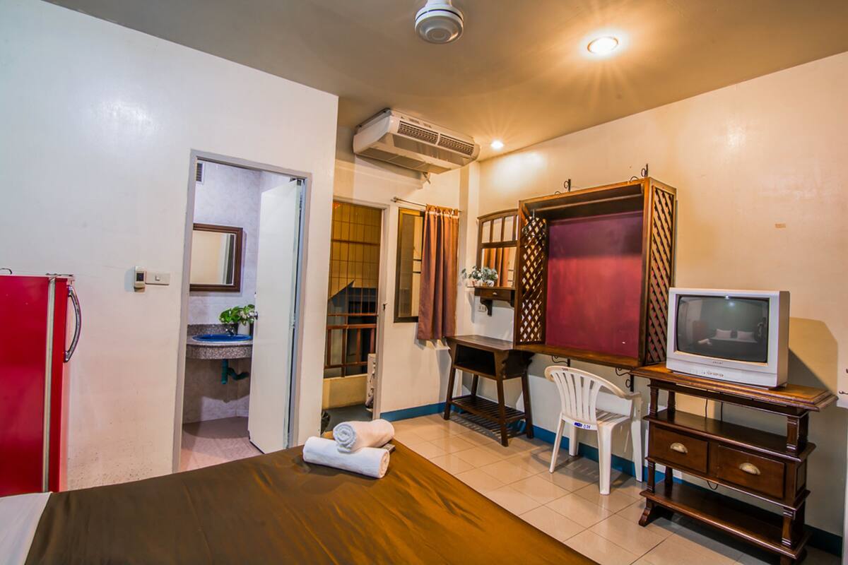 A compact hotel room with a bed, a desk with a television, a chair, a small bathroom with a sink, and an air conditioning unit.