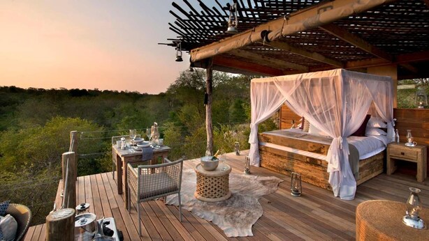 A wooden deck with a canopy bed, a dining table set with glassware, and a rustic ambiance. - Ivory Lodge (Dete)